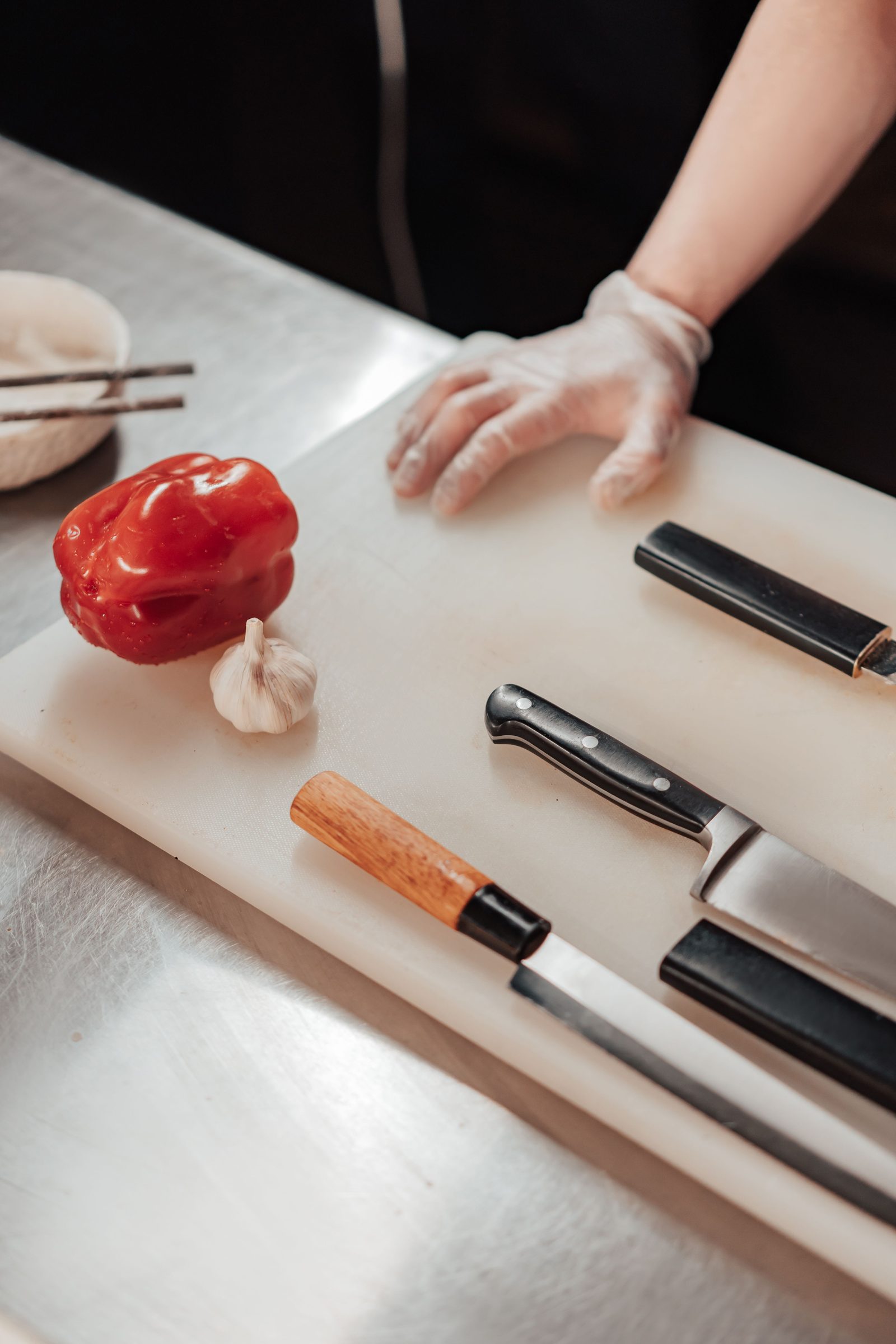 Behandschuhte Hand eines Kochs neben Kochmessern, Paprika und Knoblauch auf einem Schneidbrett in der Gewerbeküche