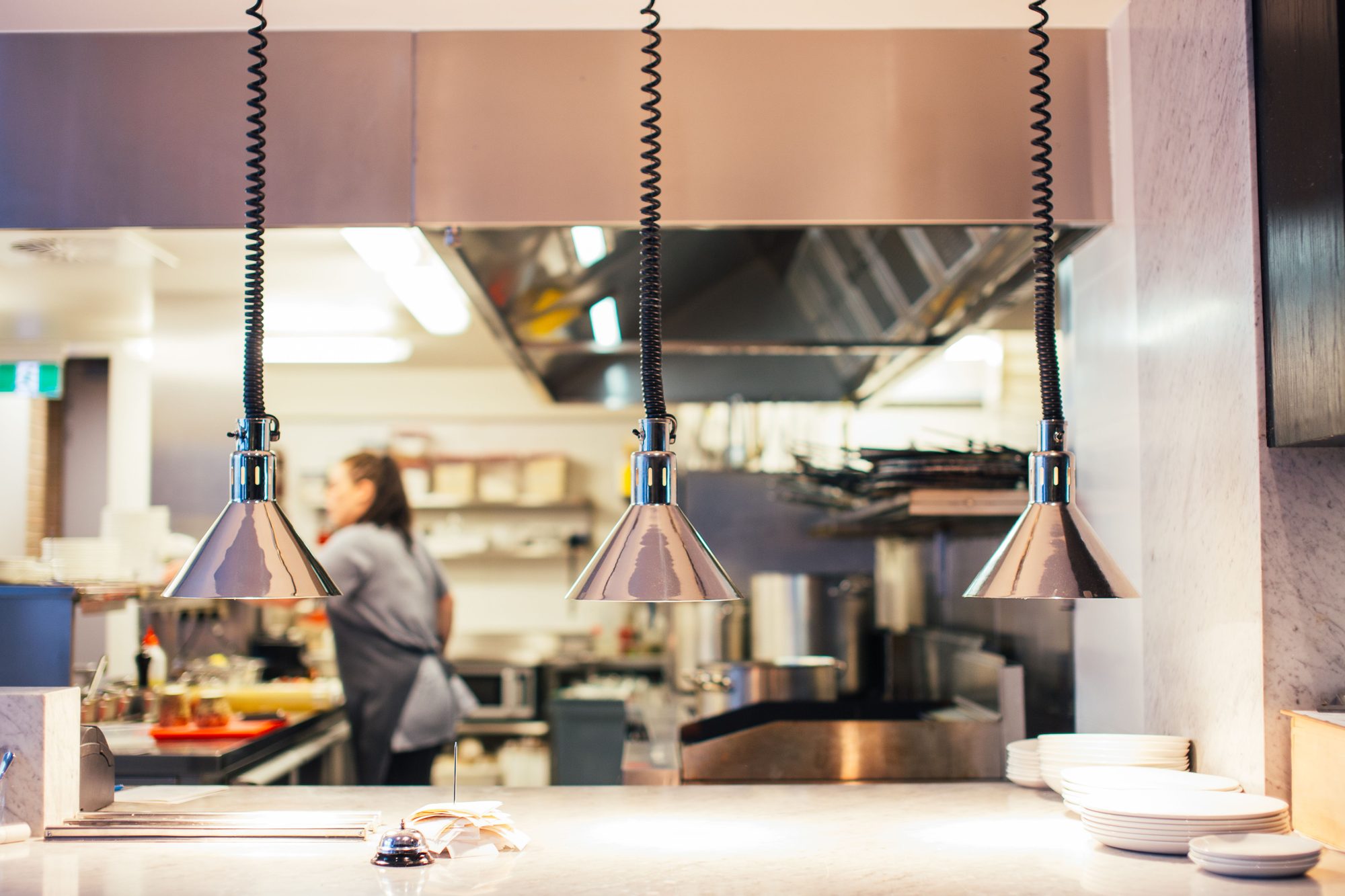 Anrichtestation einer Gastronomieküche mit Wärmelampen über der Ausgabetheke, im Hintergrund das Küchenteam bei der Arbeit